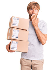 Handsome caucasian man with beard holding delivery packages covering mouth with hand, shocked and...
