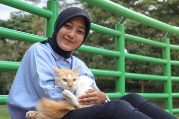 Outdoor portrait of Asian hijab woman holding and giving gentle touch to cat, taking care of her pet in nature park. Love relationship between humans and animals.