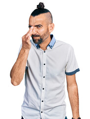 Hispanic man with ponytail wearing casual white shirt smelling something stinky and disgusting, intolerable smell, holding breath with fingers on nose. bad smell
