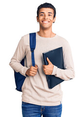 Young african amercian man wearing student backpack holding binder looking positive and happy standing and smiling with a confident smile showing teeth