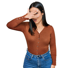 Young hispanic woman wearing casual clothes peeking in shock covering face and eyes with hand, looking through fingers with embarrassed expression.