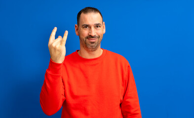 Hispanic man with beard in his 40s wearing a red sweatshirt doing rock gesture on blue studio background