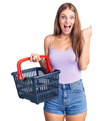 Young beautiful blonde woman holding supermarket shopping basket screaming proud, celebrating victory and success very excited with raised arms