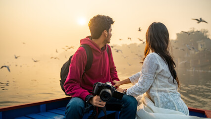 Young couple looking at the sunrise or sunset while sitting on the boat, holding a camera in hand,...