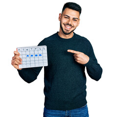 Young hispanic man with beard holding rainy weather calendar smiling happy pointing with hand and finger
