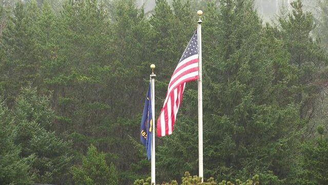 American And Oregon State Flag Flying Limply In Pouring Rain With Green Trees In Background
