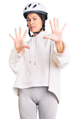 Beautiful brunette young woman wearing bike helmet and sporty clothes afraid and terrified with fear expression stop gesture with hands, shouting in shock. panic concept.