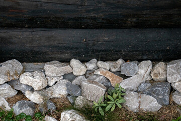 The stones lie against the log wall. The wall of the house have darkened logs with time and white stones at the base. Green grass among white stones near a log wall.