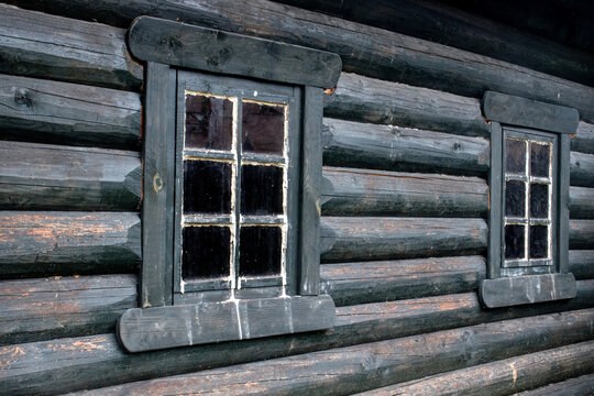 View of two windows on the wall of the log house. The wall of the house is made of old beams. Two windows on the wall of a house made of logs. The logs of the old house were darkened by age.
