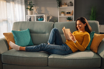 A cheerful woman is relaxing at home and reading a book. - obrazy, fototapety, plakaty