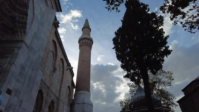 It is a historical mosque structure from the Ottoman period in Bursa. The facade of the mosque and the tree can be seen from the lower angle and in the backlight. It is moving towards the minaret sect