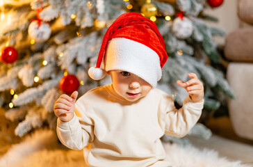 A Festive Moment: A Small Child in a Santa Hat, Standing in Front of a Christmas Tree. A small child wearing a santa hat in front of a christmas tree