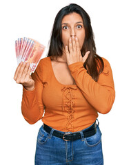 Beautiful hispanic woman holding norwegian krone banknotes covering mouth with hand, shocked and...