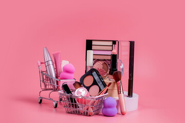 Creative concept with shopping trolley with makeup on a pink background. Perfume, sponge, brush, mascara, eye shadow, lip gloss in the basket
