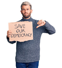 Young blond man holding save our democracy cardboard banner with angry face, negative sign showing...