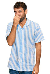 Handsome young man with beard wearing casual fresh shirt smelling something stinky and disgusting, intolerable smell, holding breath with fingers on nose. bad smell