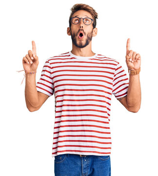 Handsome blond man with beard wearing casual clothes and glasses amazed and surprised looking up and pointing with fingers and raised arms.