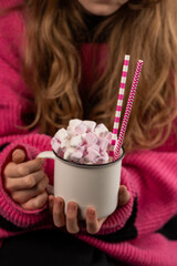 Young girl in  pink sweater is holding a mug with hot chocolate or coffee with marshmallow. Cozy holiday atmosphere, atmosphere for cold winter season. Closeup