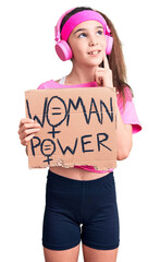 Cute hispanic child girl wearing gym clothes and using headphones holding woman power banner serious face thinking about question with hand on chin, thoughtful about confusing idea