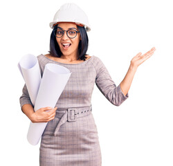 Young beautiful latin woman wearing architect hardhat and glasses holding blueprints celebrating victory with happy smile and winner expression with raised hands