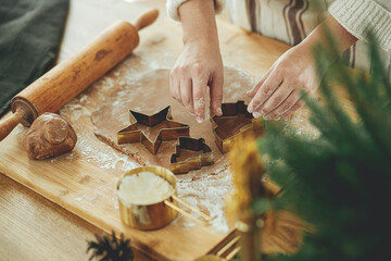 Hands cutting gingerbread dough with festive golden cutters on rustic table with holiday...