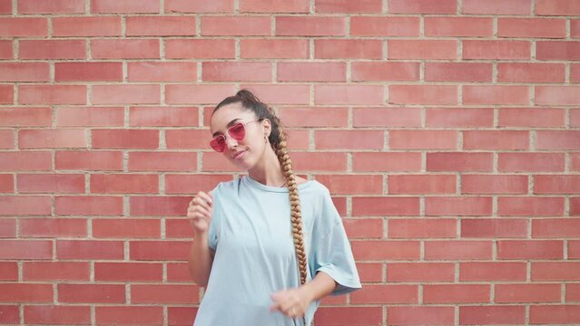 Cheerful young woman wearing sunglasses in a heart design against a brick wall in an urban setting. Stylish young woman performing fluid dance moves while smiling at the camera
