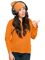 Beautiful brunette young woman listening to music using headphones smiling with happy face looking and pointing to the side with thumb up.