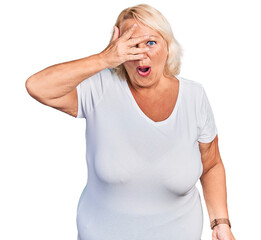 Middle age blonde woman wearing casual white t shirt peeking in shock covering face and eyes with hand, looking through fingers with embarrassed expression.