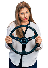 Middle age latin woman holding steering wheel in shock face, looking skeptical and sarcastic, surprised with open mouth