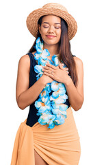 Young beautiful latin girl wearing hawaiian lei and summer hat smiling with hands on chest with...