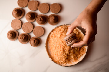 Confectioner's hand holds a brown macaroon over a plate with crumbs for decoration