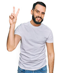 Naklejka premium Young man with beard wearing casual white t shirt smiling looking to the camera showing fingers doing victory sign. number two.