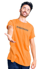Young hispanic man wearing t shirt with happiness word message smiling cheerful offering palm hand...