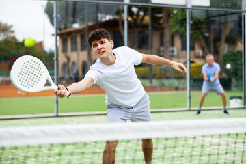 Young European male player serving ball during training padel in court outdoors in spring