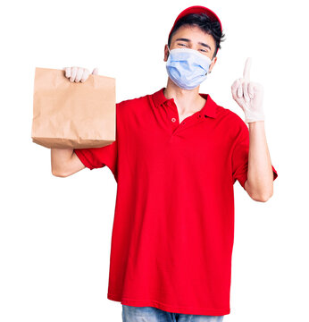 Young hispanic man wearing delivery uniform and medical mask holding paper bag surprised with an idea or question pointing finger with happy face, number one