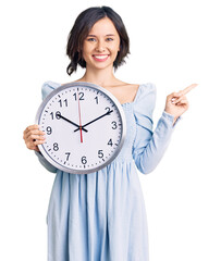 Young beautiful girl holding big clock smiling happy pointing with hand and finger to the side