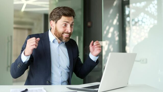 Excited happy businessman in formal suit received great news on laptop sitting at workplace in business office. Joyful entrepreneur reads a pleasant message, celebrates success, rejoices at a surprise