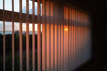 Shallow focus on vertical window blinds with a golden glow as the sun sets.There is a view of the sea and the glow of the setting sun. - 687717161