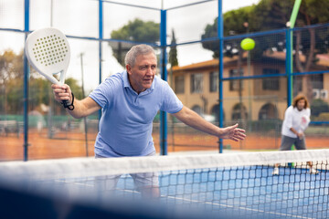 Focused aged man playing friendly paddleball match on outdoor summer court. Senior people sports...
