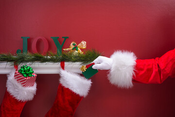Santa Claus placing two gift cards into Christmas stocking hanging from mantle