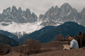 Young Attractive Man With Short-Cut Beard Taking A Well-Deserved Rest And Having A Delicious Meal After Exhausting Mountain Hike In Front Of Massive Alpine Rocks In The Dolomites Covered With Snow