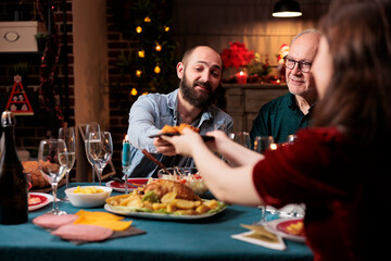 Persons eating food at festive dinner, celebrating christmas eve together and passing meal plates around. Joyful group of people enjoying holiday celebration event at home, winter feast.