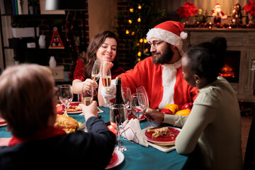 Young man santa claus having fun with friends at dinner, enjoying christmas holiday spirit together at table. People celebrating seasonal event with food and alcohol, gathering to tell stories.