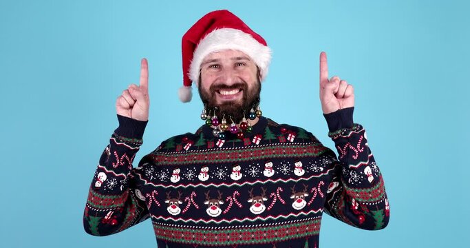 Merry bearded man wear knitted christmas sweater Santa hat posing point index finger above indicate on workspace area isolated on blue background Happy New Year 2024 celebration holiday concept. 