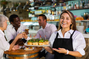 Cheerful smiling female waiter holding served tray meeting visitors at comfortable bar