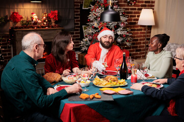 Man in santa claus costume sitting at table with diverse people, enjoying festive dinner on christmas eve festivity. Young person in suit spreading positivity during december holidays.