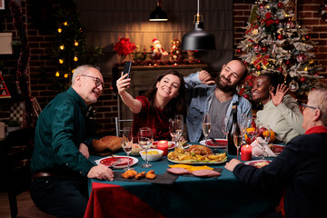 Family taking photos at christmas dinner with delicious food and glasses of wine, making memories during december holiday event. Diverse people having fun with pictures on smartphone at home.