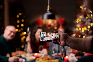 People posing for pictures at home, enjoying festive dinner feast with glasses of wine surrounded by decorations. Diverse persons taking photos during christmas eve holiday celebration.