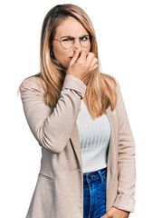 Young blonde woman wearing business shirt and glasses smelling something stinky and disgusting, intolerable smell, holding breath with fingers on nose. bad smell