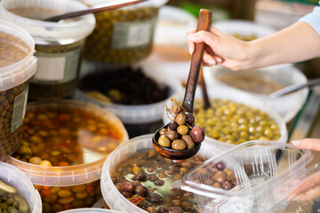 In self-service store, womens hands hold ladle and place pickled black olives with stone in...